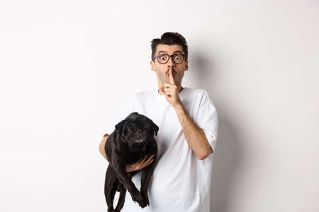 Photo pour Image of funny young man holding cute black pug dog and shushing at camera, showing hush sign and staring at camera, keeping a secret, standing over white background - image libre de droit