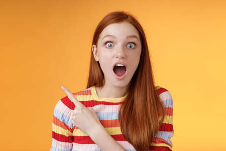 Impressed speechless young redhead 20s girl drop jaw gasping amused wide eyes staring camera surprised see awesome stunning new product pointing left index finger questioned, orange backgroundの写真素材