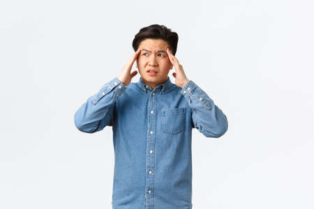 Troubled and uneasy asian man looking perplexed, having complicated situation. Guy with headache touching head and looking away, suffering painful migraine, standing white backgroundの写真素材