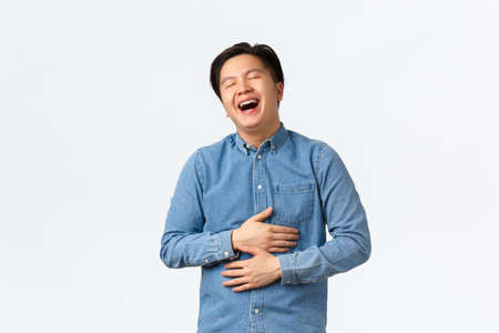 Carefree joyful handsome asian male student having fun, watching comedy, laughing out loud with closed eyes. Guy chuckle and touching belly, enjoying hilarious movie, white backgroundの写真素材