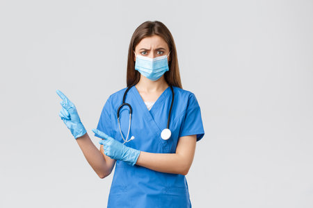 Preventing virus, health, healthcare workers and quarantine concept. Angry concerned female nurse in blue scrubs, doctor wearing medical mask, frowning and pointing left at bad infoの写真素材