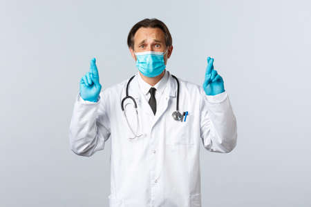 Covid-19, preventing virus, healthcare workers and vaccination concept. Troubled hopeful doctor in medical mask and gloves, cross fingers good luck, praying or making wish, white backgroundの写真素材