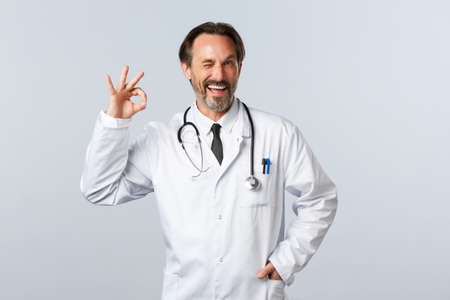 Covid-19, coronavirus outbreak, healthcare workers and pandemic concept. Cheerful happy doctor in white coat assure or recommend medicine, showing okay guarantee gesture and winkの写真素材