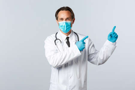 Covid-19, preventing virus, healthcare workers and vaccination concept. Smiling friendly doctor in white coat, medical mask and gloves pointing top right advertisement, white backgroundの写真素材