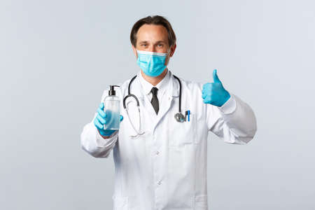 Covid-19, preventing virus, healthcare workers and vaccination concept. Satisfied doctor in medical mask and gloves thumb-up in approval, recommend hand sanitizer, white backgroundの写真素材