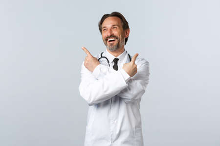 Covid-19, coronavirus outbreak, healthcare workers and pandemic concept. Carefree cheerful male doctor laughing over promo at top left. Therapist in white coat pointing sideways at advertisementの写真素材