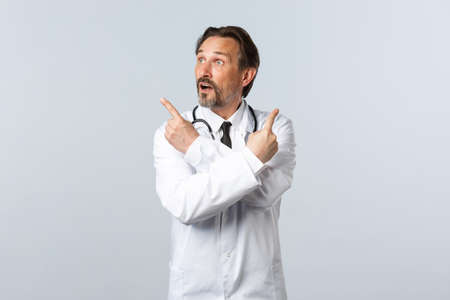 Covid-19, coronavirus outbreak, healthcare workers and pandemic concept. Unsure indecisive doctor in white coat, pointing fingers sideways and look left at variant, making choiceの写真素材