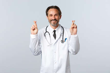 Covid-19, coronavirus outbreak, healthcare workers and pandemic concept. Optimistic smiling doctor having faith in clinic service quality, cross fingers good luck, making wishの写真素材