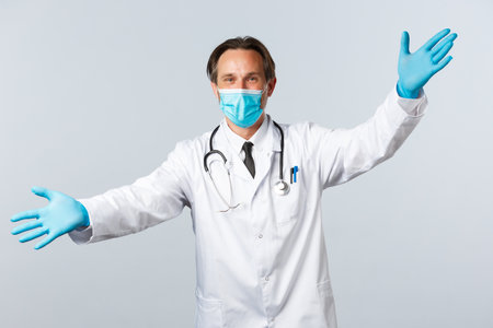 Covid-19, preventing virus, healthcare workers and vaccination concept. Friendly doctor in medical mask and gloves spread hands welcoming, greeting clinic patients, inviting come inの写真素材