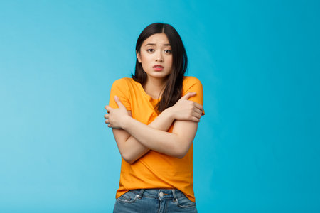 Girl feeling uncomfortable walking light yellow t-shirt, hugging herself trembling, shaking feeling cold, freezing windy weather, frowning grimacing discomfort, stand blue backgroundの写真素材