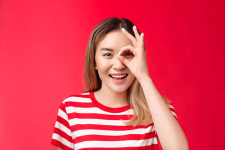 Close-up upbeat positive asian girl look through ok sign smiling broadly, enjoy positive vibes, judging, grinning spend great summer holidays, approve cool idea, stand red backgroundの写真素材