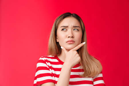 Close-up suspicious thoughtful girlfriend pondering, touch chin squinting doubtful and frowning, look up while thinking, take decision, stand red background perplexed, hesitating making choiceの写真素材