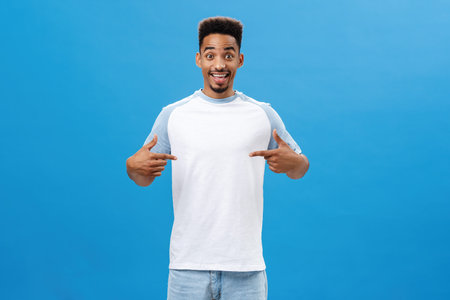 Charming dark-skinned guy asking advice what wear on date pointing at t-shirt with cheerful happy...