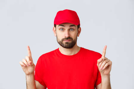 Online shopping, delivery during quarantine and takeaway concept. Indecisive and unsure courier in red uniform cap, t-shirt, smirk and pointing fingers up uncertain, dont know, cant decideの写真素材