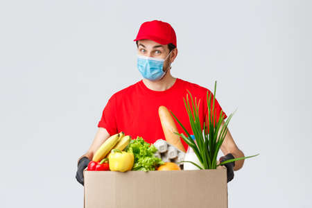 Friendly courier in face mask and gloves, red uniform bring food box to customer ordered online, contactless deliverの写真素材