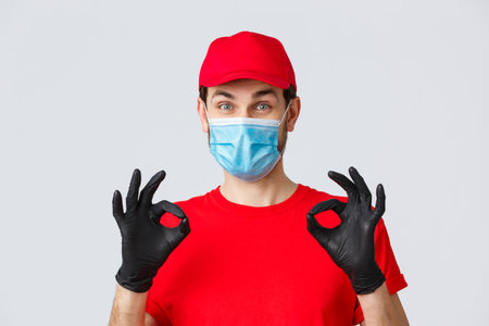 Cheerful, pleased courier in red uniform, cap and medical mask with gloves show okay, approval or...
