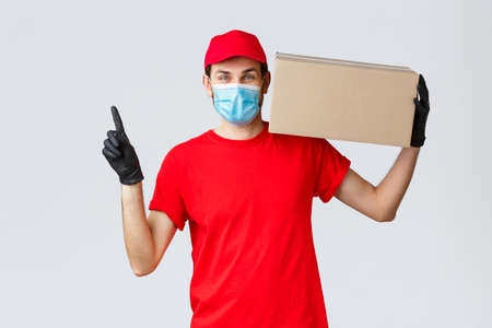 Cheerful courier in face mask, gloves and uniform, carry your parcel to doorsteps, pointing up, hold box with orderの写真素材