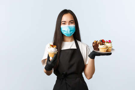 Friendly cute asian barista, worker in medical mask handing you muffin, serving order in cafeの写真素材