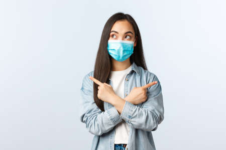 Thoughtful indecisive asian girl making choice between two variants, pointing sideways and look left, wear maskの写真素材