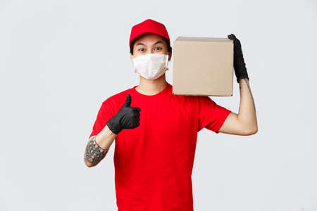 Concept of delivery and carriers during coronavirus pandemic. Smiling asian courier in red cap, t-shirt, wearing medical mask and gloves, covid-19 safety measures on work, hold package, make thumb-upの写真素材