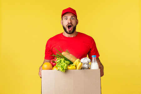 Online delivery, shopping and food order concept. Excited and amused courier in red uniform, bearde delivery man gasping astonished, bring grocery box to customer home, yellow backgroundの写真素材