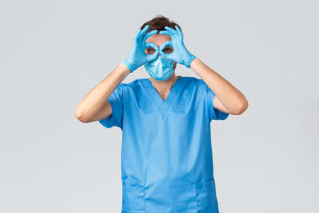 Coronavirus outbreak, healthcare workers fighting disease, hospitals concept. Funny male doctor in scrubs and medical mask, gloves showing goggles, glasses with hands over eyes, fool around on lunchの写真素材