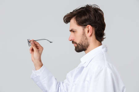 Hospital, healthcare workers, covid-19 treatment concept. Profile portrait of serious-looking doctor in scrubs, looking at glasses, examine patient eyewear, physician picking eyewear for patientの写真素材