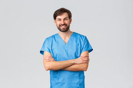 Healthcare workers, coronavirus quarantine campaign and pandemic concept. Smiling bearded caucasian male doctor or nurse in blue scrubs, cross hands chest and laughing, have break in hospitalの写真素材