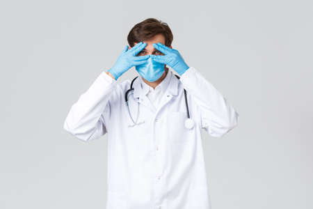 Hospital, healthcare workers, covid-19 treatment concept. Handsome doctor in medical mask, scrubs and gloves, looking through fingers, peeking at camera, standing grey backgroundの写真素材