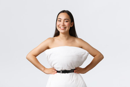 Beautiful asian girl making dress from pillow, secure it with belt around waste and posing for social media, smiling pleased, standing white background delightedの写真素材