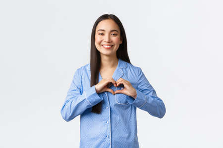 Cute tender asian girl in blue jammies showing heart gesture and smiling, express love and care, confess sympathy, standing in pajamas liking awesome sleepover, white backgroundの写真素材