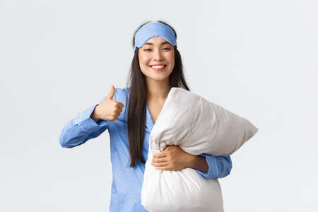 Cheerful pleased cute asian girl in blue pajama and sleeping mask, holding soft comfortable pillow and showing thumbs-up satisfied, had good night sleep, taking pills from insomniaの写真素材