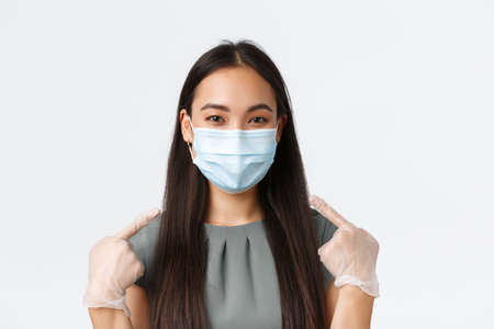 Small business owners, covid-19, preventing virus measures concept. Close-up of smiling asian woman in medical mask and gloves pointing at face, using protective equipment during pandemicの写真素材