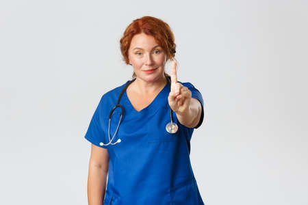 Medicine, healthcare and virus concept. Smiling female middle-aged doctor, medical worker in scrubs shaking finger in warning or disapproval, prohibit something, restrain from actionの写真素材