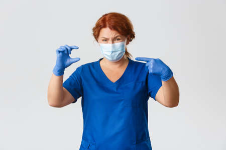 Medical workers, pandemic, virus concept. Upset and disappointed female doctor, middle-aged nurse in face mask, rubber gloves pointing at something small or little with sad expressionの写真素材