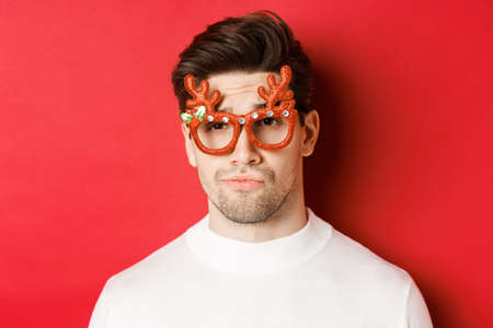 Concept of winter holidays, christmas and celebration. Close-up of skeptical handsome guy, wearing party glasses, smirk and looking unamused, standing over red backgroundの写真素材