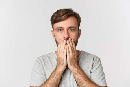 Close-up of shocked and speechless guy, cover mouth and looking at camera, standing over white backgroundの写真素材