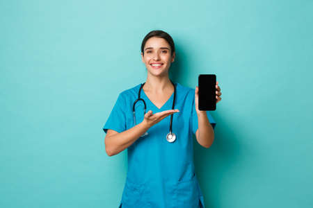 Pandemic and medicine concept. Beautiful female doctor, smiling and demonstrating mobile phone application, showing online check up app, standing over blue backgroundの写真素材