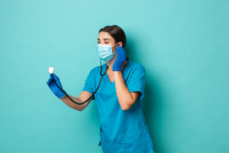 Pandemic and medicine concept. Image of beautiful female doctor in medical mask, scrubs and gloves, checking patient with stethoscope, looking left, standing over blue backgroundの写真素材