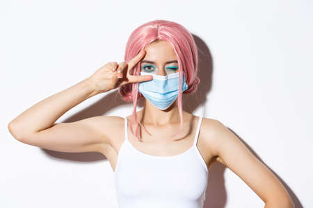 virus, social distancing and lifestyle concept. Close-up of beautiful cheerful girl in medical mask and pink wig, staying positive, showing peace sign and winking joyfullyの写真素材