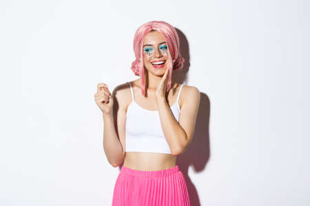 Image of pleased stylish girl in pink wig looking happy at credit card, dreaming of shopping, standing over white backgroundの写真素材