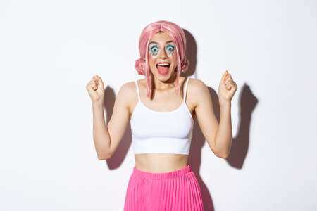 Image of cheerful girl in pink wig and halloween costume, raising hands up like a winner, celebrating victory, looking amazed and delighted, standing over white backgroundの写真素材