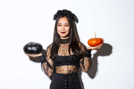 Pleased smiling asian woman celebrating halloween, wearing witch costume, holding pumpkins, standing over white backgroundの写真素材