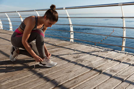 Young attractive fitness woman tying her shoes, jogging on the seaside promenadeの写真素材