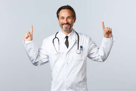 virus outbreak, healthcare workers and pandemic concept. Cheerful smiling male doctor in white coat, pointing fingers up to show banner, promote product medicationの写真素材
