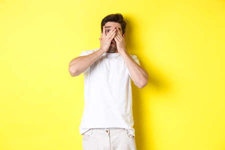 Embarrassed guy shut eyes but peeking through fingers at something awkward, standing over yellow backgroundの写真素材
