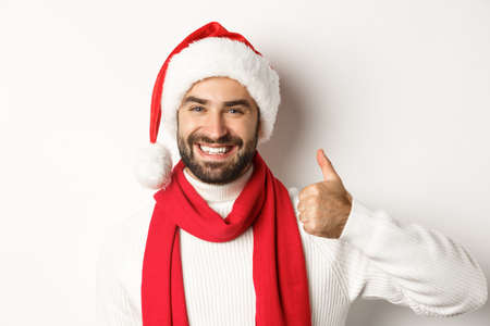 New Year party and winter holidays concept. Close-up of happy man in Santa hat showing thumbs up in approval, like and agree, standing over white backgroundの写真素材
