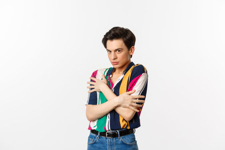 Image of timid gay man cringe and looking displeased at camera, hugging himself defensive, standing...
