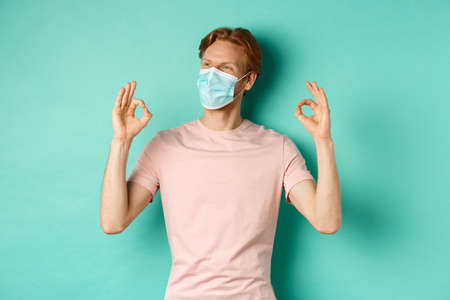 Covid-19, pandemic and lifestyle concept. Cheerful redhead man in medical mask showing okay signs in approval, like and praise product, looking left at copy spaceの写真素材