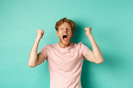 Handsome redhead man watching sports game and cheering, shouting and making fist pumps, looking at...